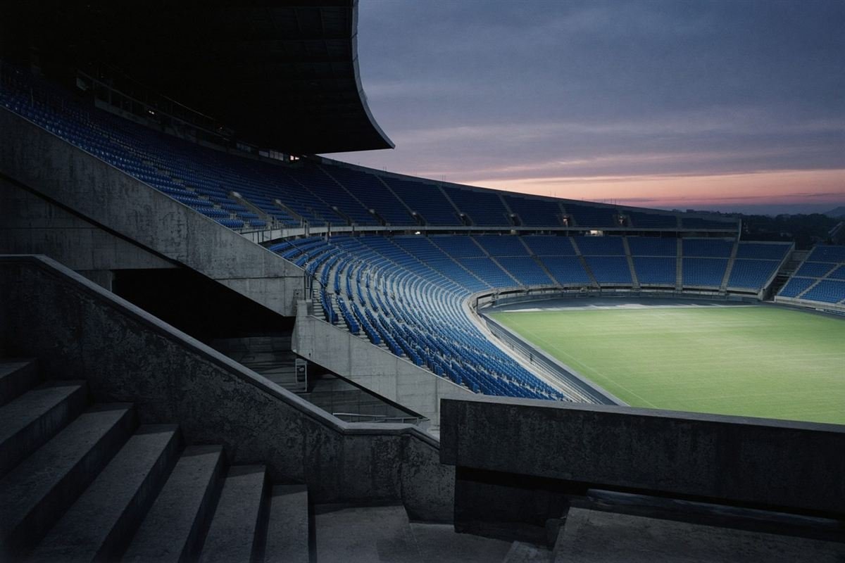 An empty stadium at twilight, cobalt seats around a floodlit pitch.
